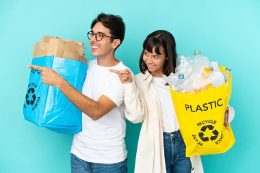 Mavi arka planda izole edilmiş plastik ve kâğıt dolu bir çanta taşıyan genç çift, bir ürünü sunmak için yan tarafı işaret ediyor.