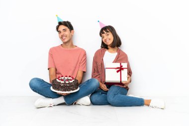 Genç melez çift, beyaz arka planda izole bir şekilde yerde oturmuş, doğum gününü kutlarken kollarını yan yana ve gülümseyerek geçiriyorlar.