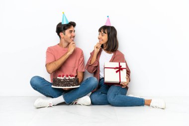 Genç melez bir çift, doğum gününü kutluyorlar. Beyaz arka planda izole bir şekilde yerde oturmuş birbirlerine bakıyorlar.