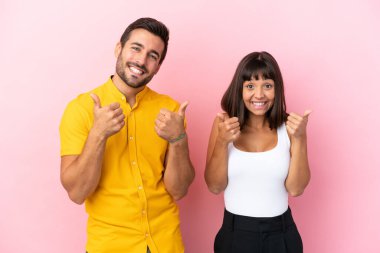 Pembe arka planda izole edilmiş genç bir çift iki eliyle baş parmak işareti yapıyor ve gülümsüyor.
