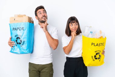 Geri dönüşüm için plastik ve kağıt dolu bir çanta taşıyan genç çift beyaz arka planda izole edilmiş bir şekilde yukarı bakarken bir fikir düşünüyor.