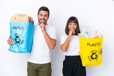 Beyaz arka planda izole edilmiş plastik ve kâğıt dolu bir çanta taşıyan genç çift tatlı bir ifadeyle gülümsüyor.