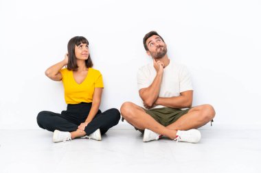 Genç bir çift beyaz arka planda izole bir şekilde yerde oturuyor. Kafalarını kaşıyarak bir fikir üretiyorlar.