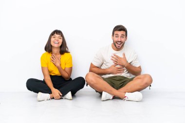 Yerde oturan genç çift, beyaz arka planda soyutlanmış bir şekilde gülümserken ellerini göğsüne koyuyor.