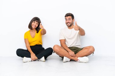 Genç çift, beyaz arka planda izole bir şekilde yerde oturuyor ve iyi bir şey olduğu için iyi bir jest yapıyorlar.