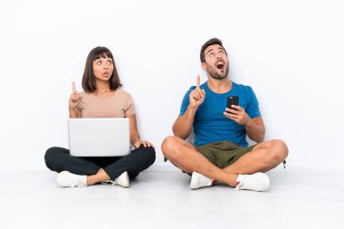 Yerde oturan genç çift beyaz arka planda izole edilmiş cep telefonu ve cep telefonlarını tutarken bir fikir öne sürüyorlar.