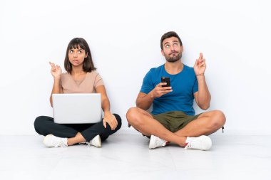 Yerde oturan genç çift beyaz arka planda izole edilmiş bilgisayar ve cep telefonlarını tutarken şans diliyor.