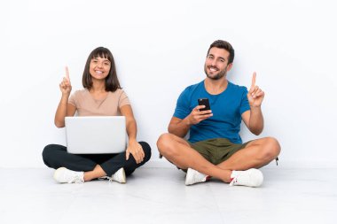 Yerde oturan genç çift beyaz arka planda izole edilmiş bilgisayar ve cep telefonlarını tutuyor ve parmak kaldırıyor.