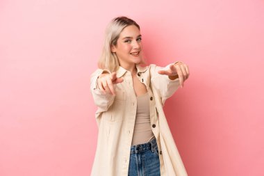 Pembe arka planda izole edilmiş genç beyaz kadın mutlu bir ifadeyle ön tarafı işaret ediyor.