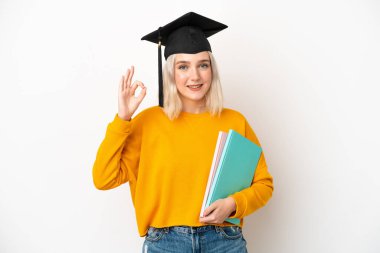 Beyaz arka planda izole edilmiş beyaz bir üniversite mezunu genç beyaz kadın parmaklarıyla tamam işareti gösteriyor.