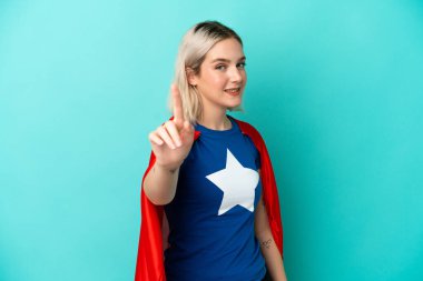 Süper kahraman beyaz kadın mavi arka planda izole edilmiş ve parmağını kaldırıyor.
