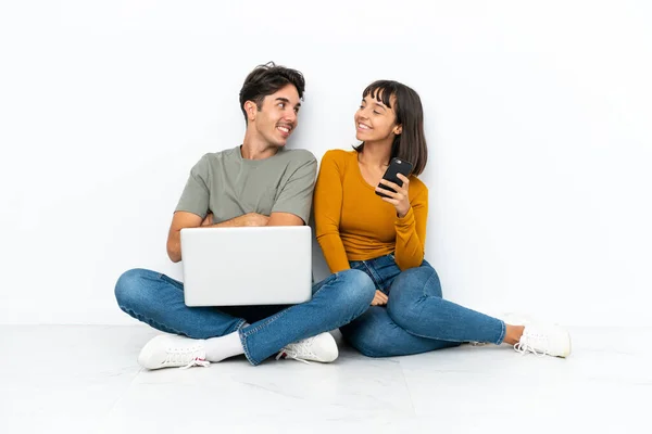 年轻的一对夫妇 带着笔记本电脑和手机坐在地板上 微笑着回头看 — 图库照片