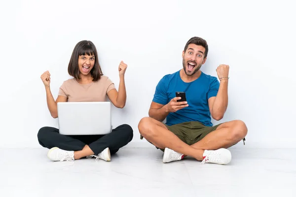 一对年轻的夫妇坐在地板上 手持白色背景的掌上电脑和手机 庆祝胜利 — 图库照片