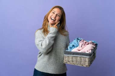 Orta yaşlı bir kadın mor arka planda izole edilmiş bir çamaşır sepetini tutarken mutlu ve gülümsüyor.