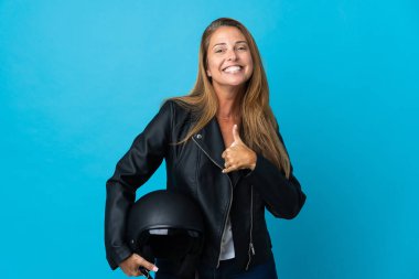 Mavi arka planda izole edilmiş motosiklet kaskı tutan orta yaşlı bir kadın baş parmak işareti yapıyor.