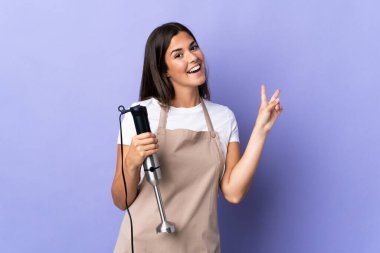 Mor arka planda izole edilmiş el blenderi kullanan Brezilyalı kadın gülümsüyor ve zafer işareti gösteriyor