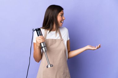 Mor arkaplanda yalıtılmış el blenderi kullanan Brezilyalı kadın yan bakarken sürpriz yüz ifadesi kullanıyor