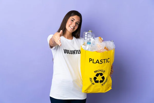 若いブラジル人女性は袋を持っていますプラスチックボトルでいっぱい紫色の背景に隔離されたリサイクル良い取引を閉じるために握手 — ストック写真