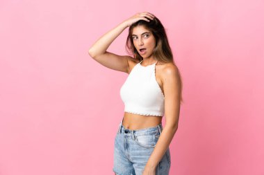 Pembe arka planda izole edilmiş genç beyaz kadın yan tarafa bakarken sürpriz jest yapıyor.