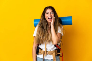 Sarı arka planda izole edilmiş büyük sırt çantalı genç dağcı kadın ağzı açık bağırıyor.