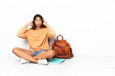 Genç bir kız öğrenci dizüstü bilgisayarla yerde oturuyor. Beyaz arka planda izole edilmiş. Şüpheleri var ve düşünüyor.