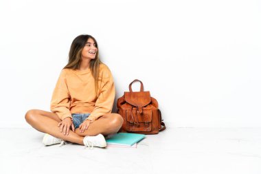 Yerde dizüstü bilgisayarlı genç bir bayan oturuyor. Beyaz arka planda izole edilmiş. Yan pozisyonda gülüyor.