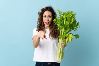 Mavi arka planda izole edilmiş bir kereviz tutan genç kadın şaşırmış ve ön tarafı işaret ediyor.