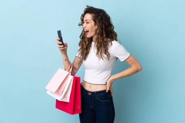 Kafkasyalı genç bir kadın mavi arka planda izole edilmiş. Elinde alışveriş poşetleri var ve bir arkadaşına cep telefonuyla bir mesaj yazıyor.