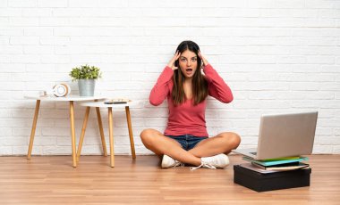 Evde dizüstü bilgisayarı olan genç bir kadın yerde oturuyor ve şaşırmış görünüyor.