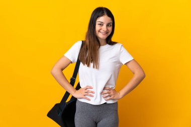 Spor çantalı genç bayan sarı arka planda izole edilmiş, kolları kalçasında ve gülümsüyor.
