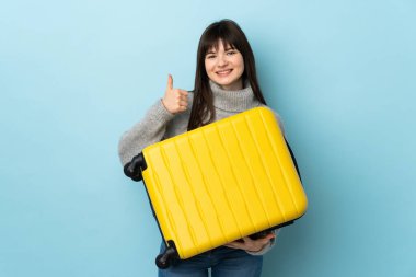 Genç Ukraynalı bir kadın mavi arka planda izole edilmiş seyahat valiziyle ve başparmağıyla