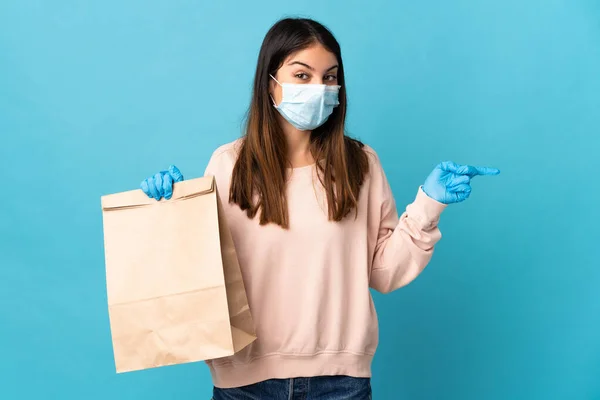 マスクでコロナウイルスから保護し 青の背景に隔離された食料品の買い物袋を持っている若い女性は驚いて 指を横に向けて — ストック写真