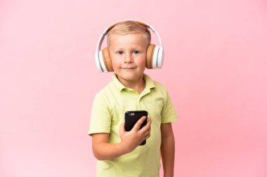 Küçük Rus çocuk pembe arka planda izole edilmiş. Cep telefonuyla müzik dinliyor ve ön tarafa bakıyor.
