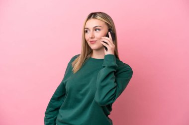 Pembe arka planda izole edilmiş genç beyaz bir kadın cep telefonuyla sohbet ediyor.