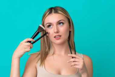 Mavi arka planda izole edilmiş beyaz bir kadın elinde makyaj fırçası tutuyor.