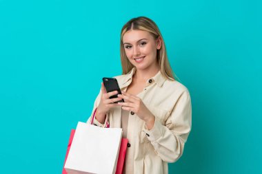Kafkasyalı genç bir kadın mavi arka planda izole edilmiş. Elinde alışveriş poşetleri var ve bir arkadaşına cep telefonuyla bir mesaj yazıyor.