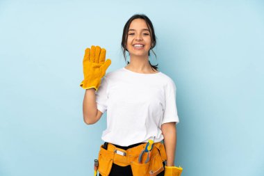 Mavi arka planda izole edilmiş genç elektrikçi kadın mutlu bir ifadeyle selamlıyor.