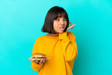 Genç esmer kadın izole edilmiş arka planda burger tutuyor hayal kırıklığına uğramış ve kulaklarını kapatmış.