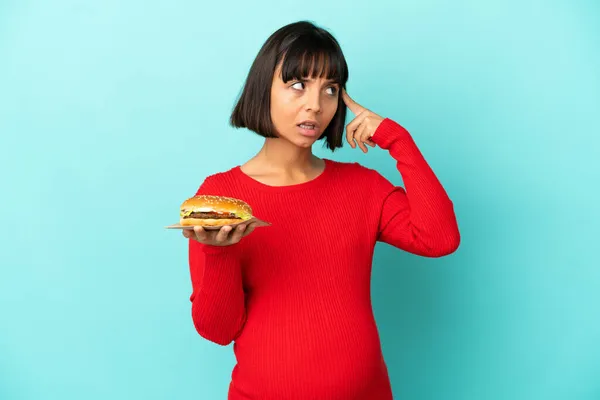 Młoda Kobieta Ciąży Trzyma Hamburgera Odosobnionym Tle Mając Wątpliwości Myślenia — Zdjęcie stockowe