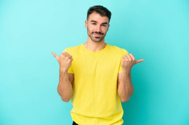 Kafkasyalı genç adam mavi arka planda izole edilmiş. Baş parmakları havada ve gülümsüyor.