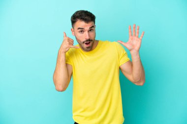Kafkasyalı genç adam mavi arka planda izole edilmiş telefon jestleri yapıyor ve şüpheli.