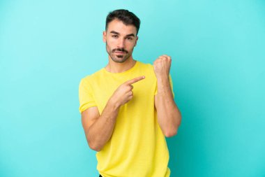 Kafkasyalı genç adam mavi arka planda izole edilmiş. Geç kalma jesti yapıyor.