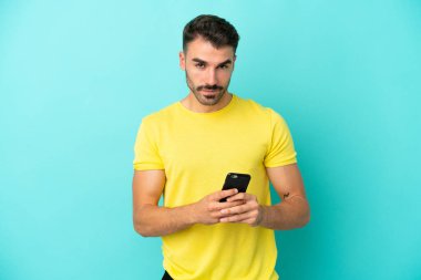 Mavi arka planda izole edilmiş genç beyaz adam cep telefonuyla bir mesaj gönderiyor.