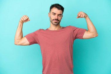 Mavi arka planda izole genç beyaz adam güçlü bir jest yapıyor.