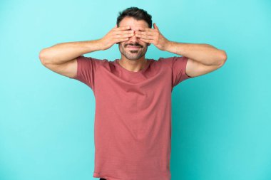 Mavi arka planda izole edilmiş genç beyaz bir adam elleriyle gözlerini kapatıyor ve gülümsüyor.