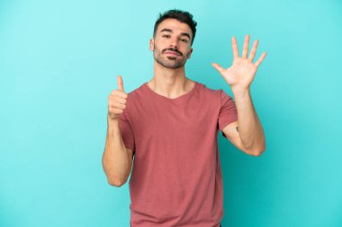 Mavi arka planda izole edilmiş genç beyaz adam parmaklarıyla altısını sayıyor.