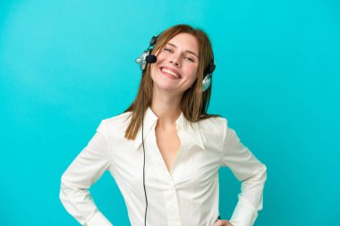 Tele pazarlamacı İngiliz kadın mavi arka planda izole edilmiş bir kulaklıkla çalışıyor. Kolları kalçasında ve gülümsüyor.