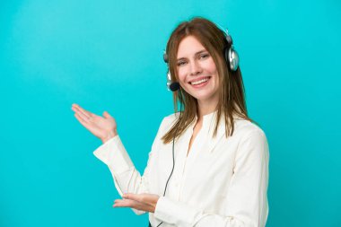 Tele pazarlamacı İngiliz kadın, mavi arka planda izole edilmiş bir kulaklıkla çalışıyor. Davet edildiği için ellerini yana uzatıyor.