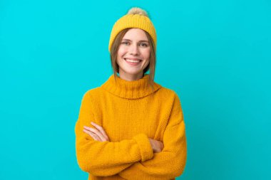 Mavi arka planda izole edilmiş kışlık ceket giyen genç İngiliz kadın kollarını ön cephede çapraz tutuyor.