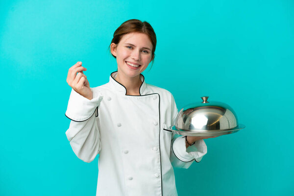 Young chef woman with tray isolated on blue background making money gesture